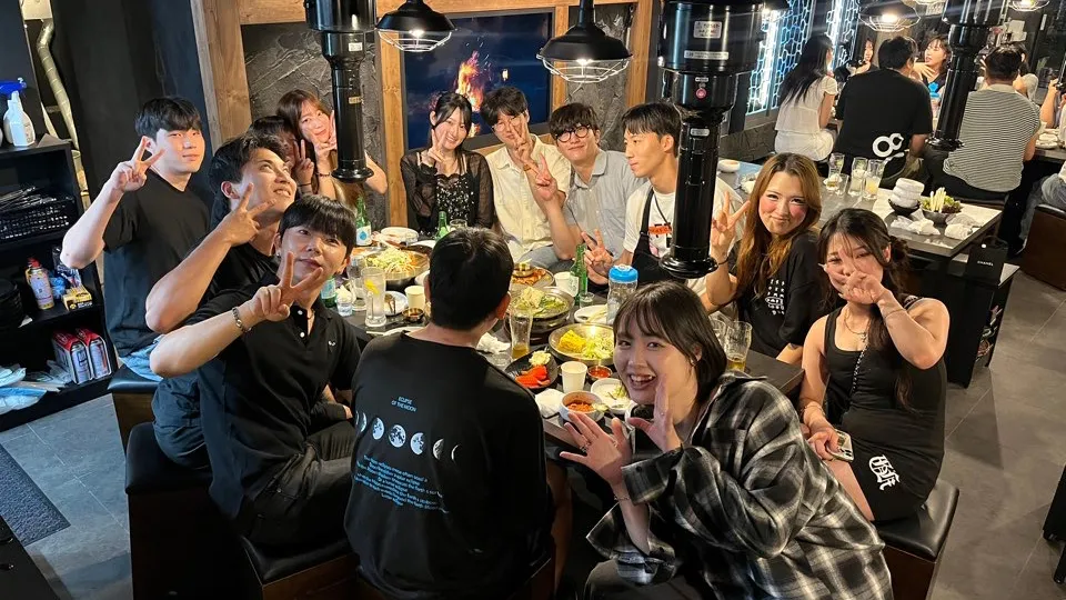 焼肉店で若い男女のグループがテーブルを囲み、料理を楽しみながら笑顔でポーズをしている。