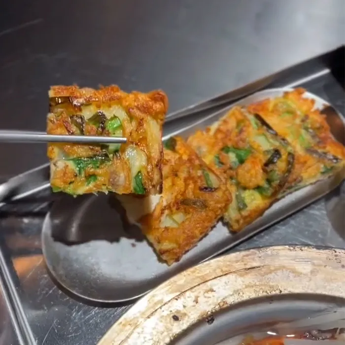 箸で持ち上げられたチヂミが金属トレイの上に並ぶほかの切れ端とともに見えます。